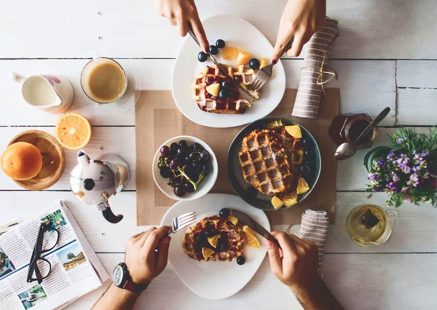 People cut up waffles at a breakfast table with coffee and fruit.