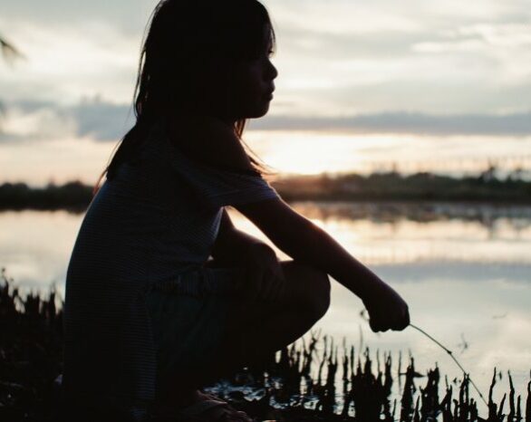 The silhouette of a child crouching along a waterfront at sunset.